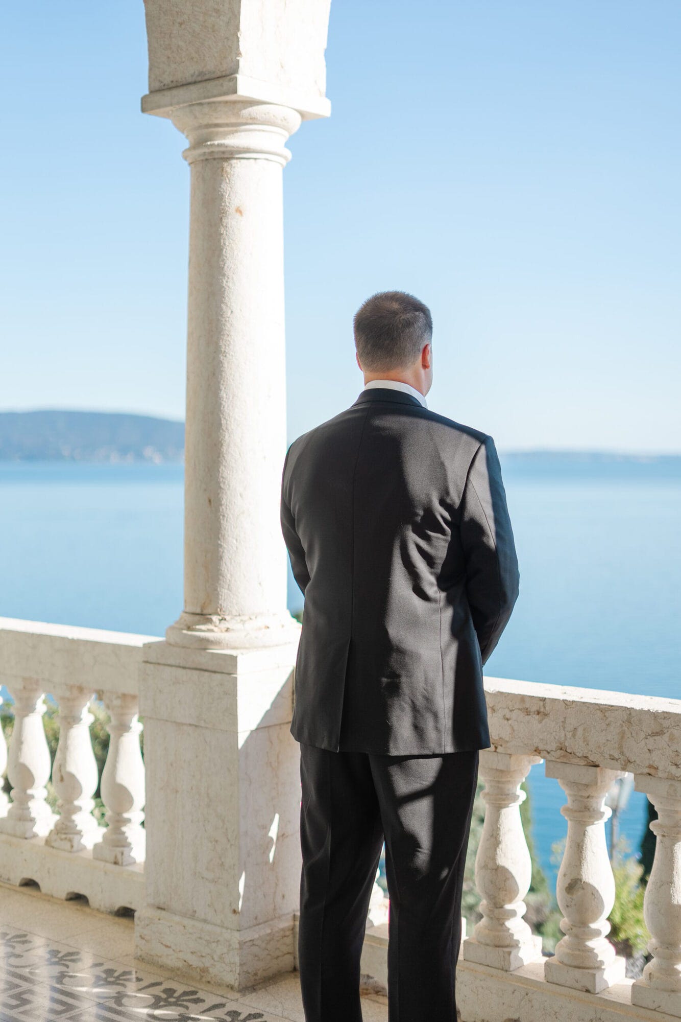 Lo sposo si trova sulla terrazza, con il Lago di Garda che fa da cornice alla scena. Di spalle, con il cuore in attesa, ogni dettaglio del suo abito impeccabile racconta la trepidante emozione prima del tanto atteso incontro con la sua futura sposa.