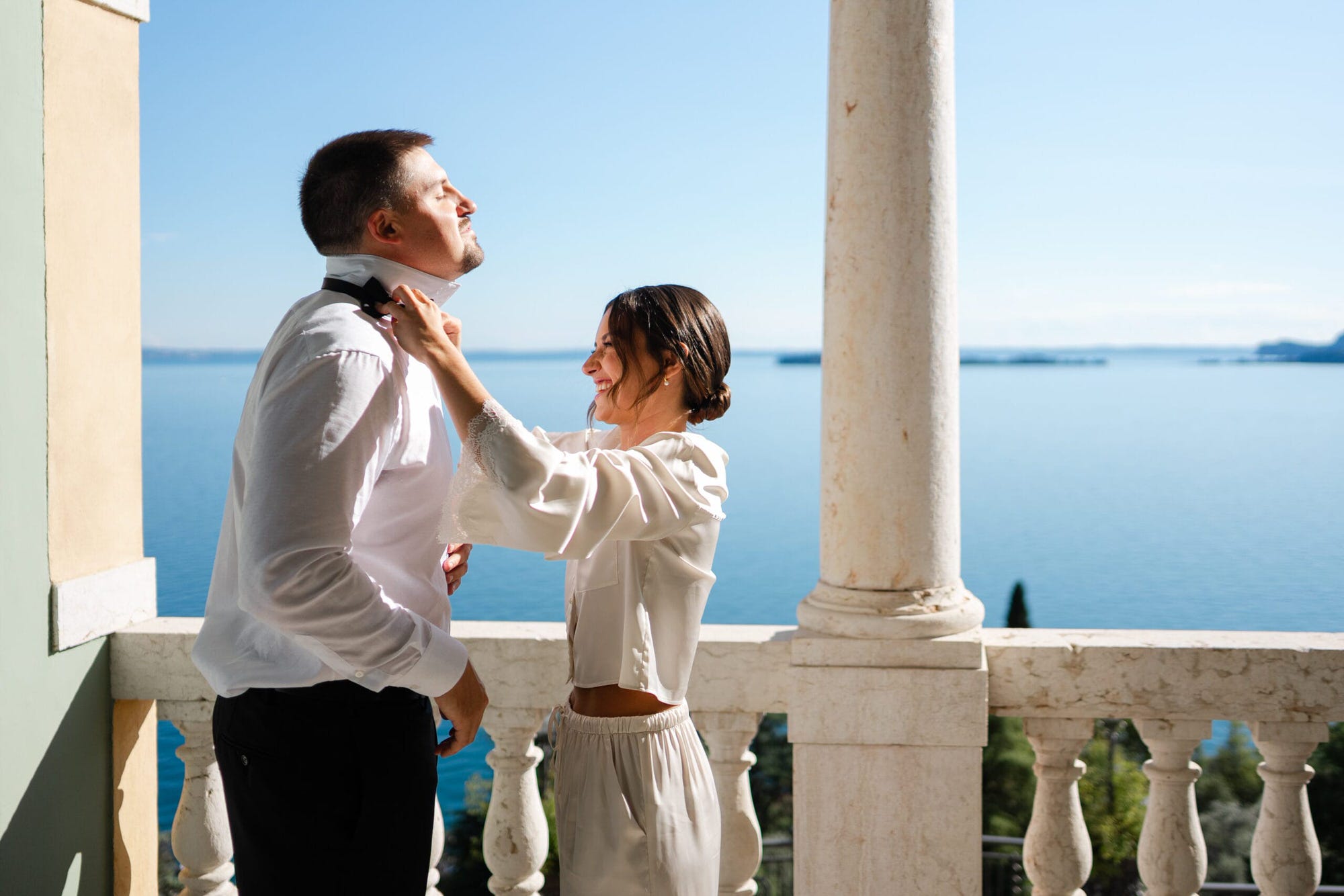 In un ambiente intimo e sereno, la sposa assiste lo sposo nei preparativi del grande giorno. La terrazza con vista sul lago crea una cornice perfetta per un gesto pieno di tenerezza, mentre i dettagli del loro abito si uniscono alla bellezza naturale che li circonda.