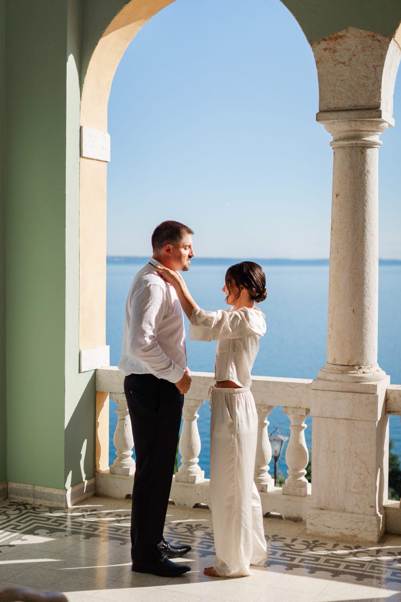 La sposa, con un sorriso tenero, aiuta lo sposo a indossare il suo abito con attenzione e amore. La terrazza che affaccia sul Lago di Garda rende questo momento ancora più speciale, creando un’atmosfera unica e carica di emozione.